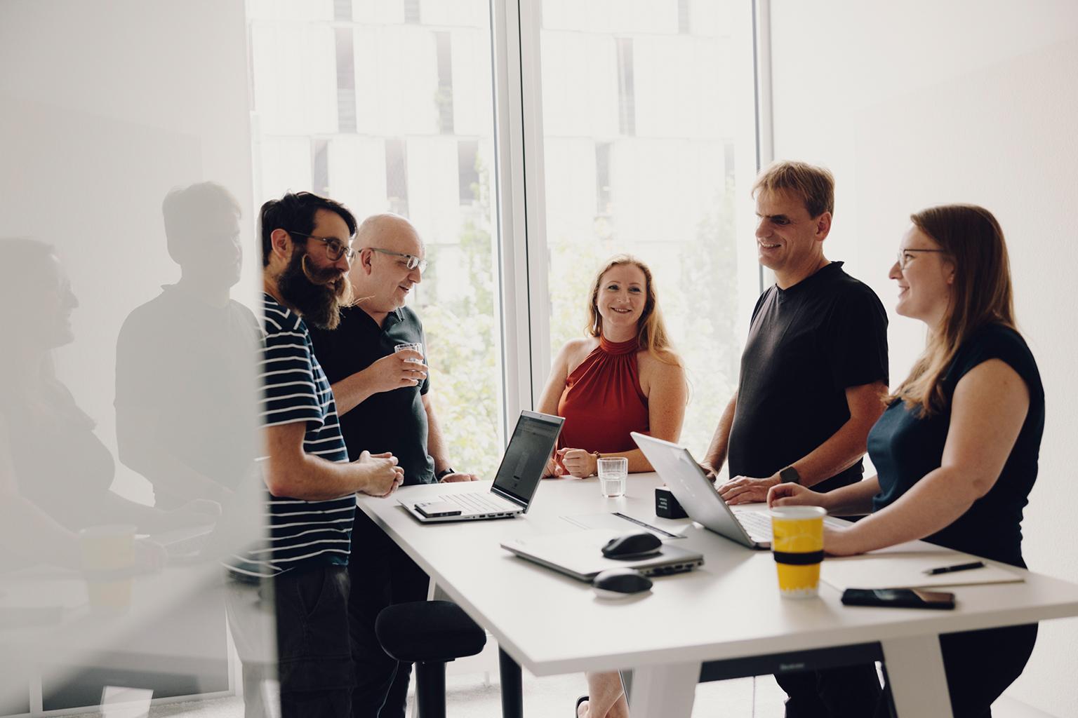 Das Bild zeigt fünf Personen in einem modernen Büroraum, die um einen Tisch stehen und diskutieren