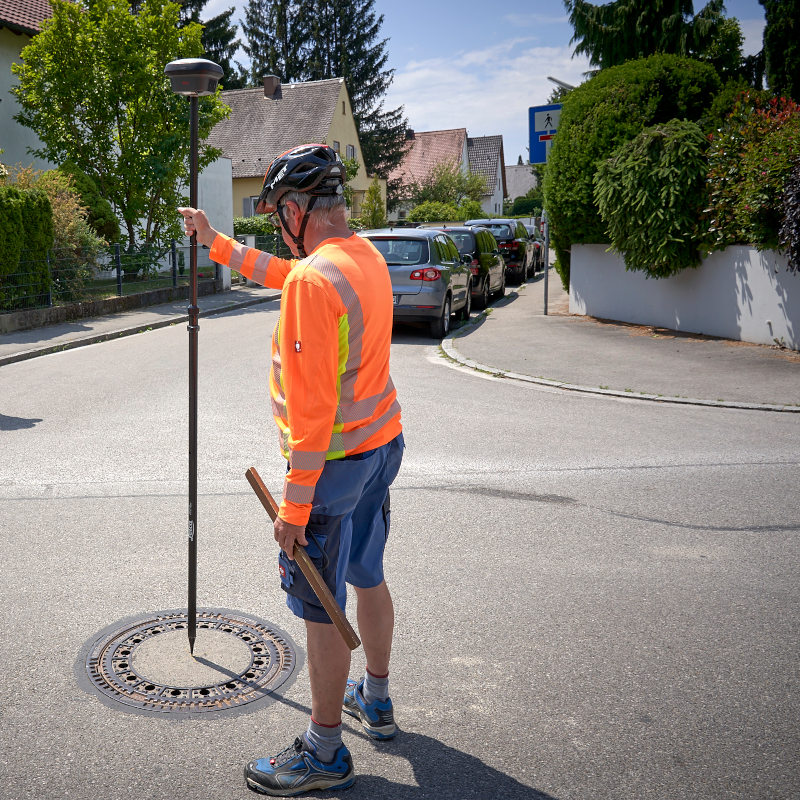 Eine Person steht auf der Straße an einem Kanaldeckel. In der Hand hält sie einen langen Stab mit GNSS-Empfänger zur Passpunktmessung.