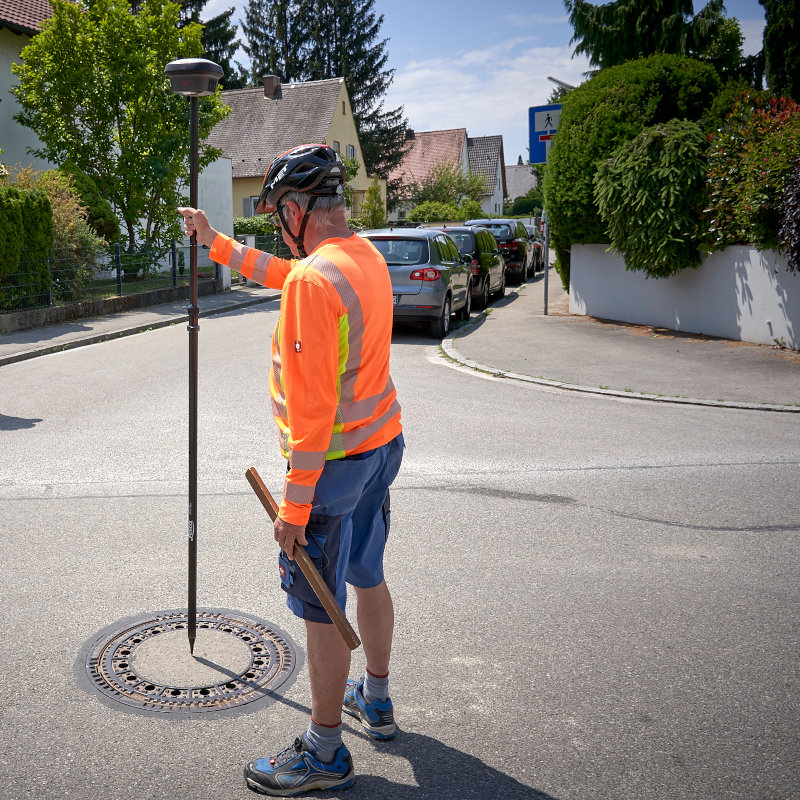 A person is standing on the street by a manhole cover. They are holding a long pole with a GNSS receiver for measuring control points.