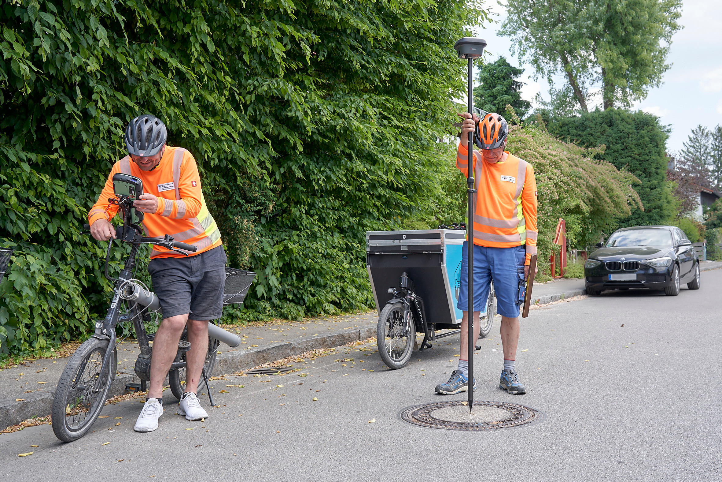 Two people in high-visibility vests on a road. One is leaning against a bicycle and holding a controller to carry out the measurements, the other is holding a GNSS receiver vertically above a manhole cover.