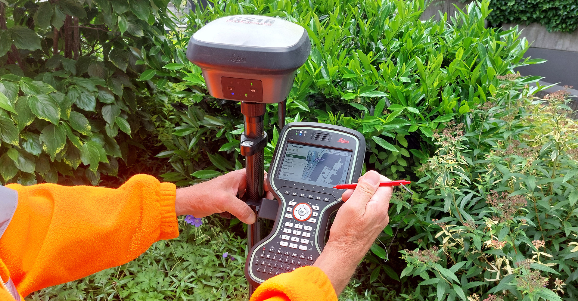 A person is holding a black stick with a plate-shaped GNSS antenna at the top. Underneath is a device with a display and keyboard on which a map is visible. The person is pointing at the display with a pen.