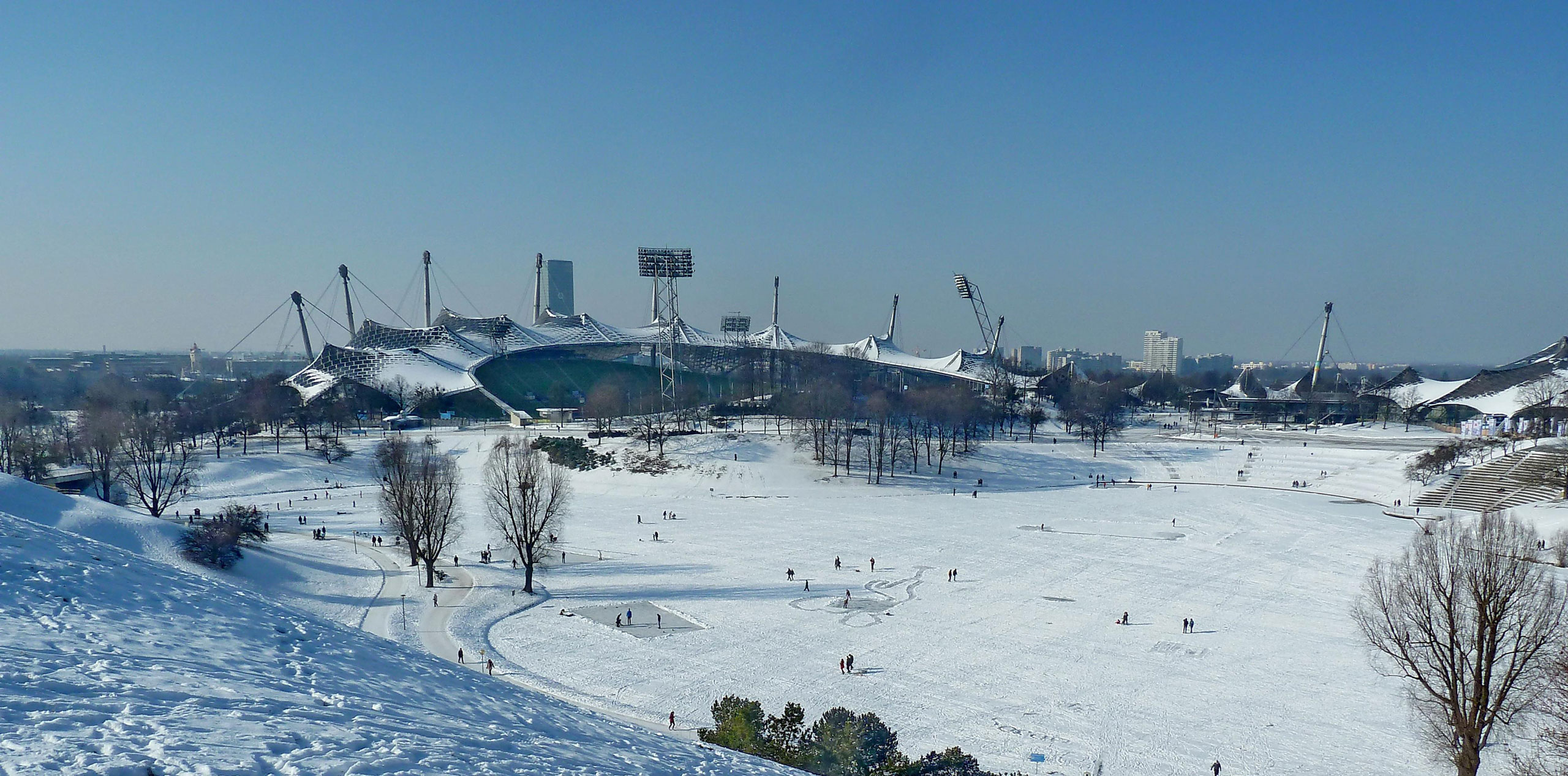 Blick auf den Olympiapark im Winter mit dem Olympiasee. Auf dem See sind Schlittschuhläufer*innen zu sehen.