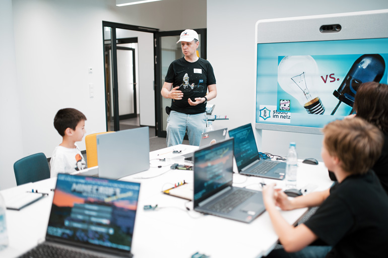 Das Foto zeigt den Workshopleiter stehend vor einigen Tischen, an denen 3 Kinder vor einem Laptop sitzen und praktische Übungen nach Anweisung des Workshopleiters ausführen.